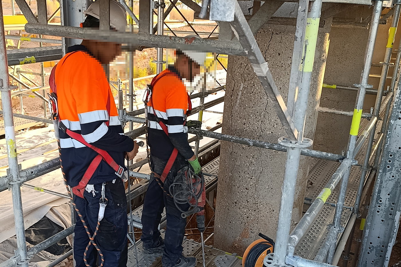 Dos operarios de Cimentaworks con casco, ropa de alta visibilidad y arnes trabajando sobre plataforma de andamio junto al soporte de hormigon